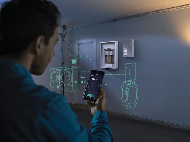A person views a smartphone displaying an energy management app. Green holographic outlines of a lightbulb, washing machine, and EV charger appear on the wall, connected to the app by dashed lines, illustrating smart home energy control.