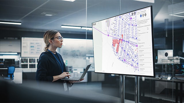 A professional woman wearing glasses stands in front of a large monitor holding a laptop.