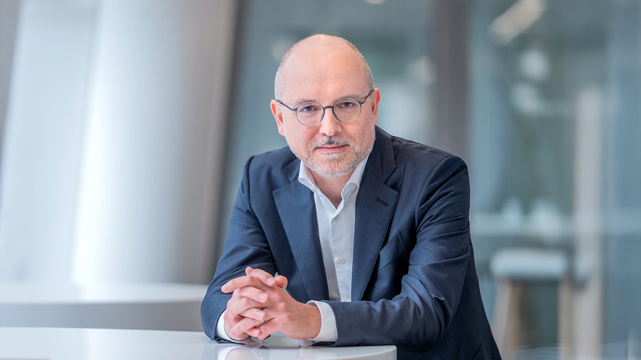 A man in a suit and tie is standing in front of a Siemens building, smiling and looking at the camera.