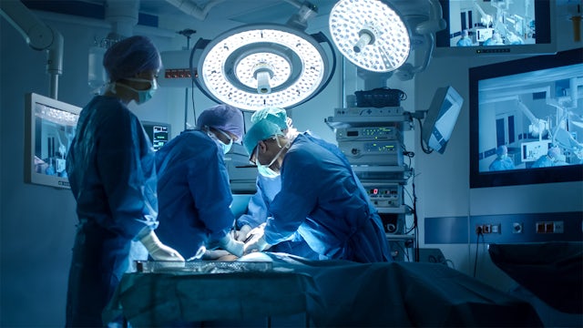 Three surgeons operate on a patient under bright lights, with a nurse standing by.