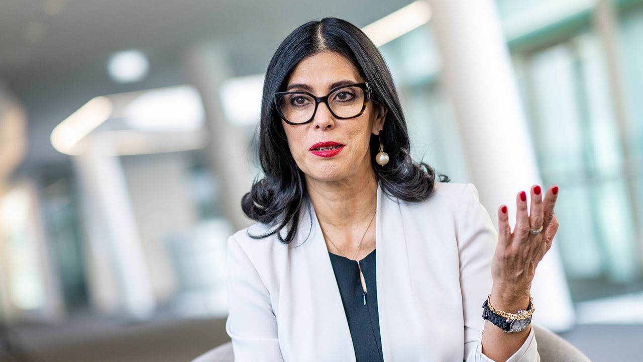 A woman in a black dress stands in front of a white wall, holding a microphone.