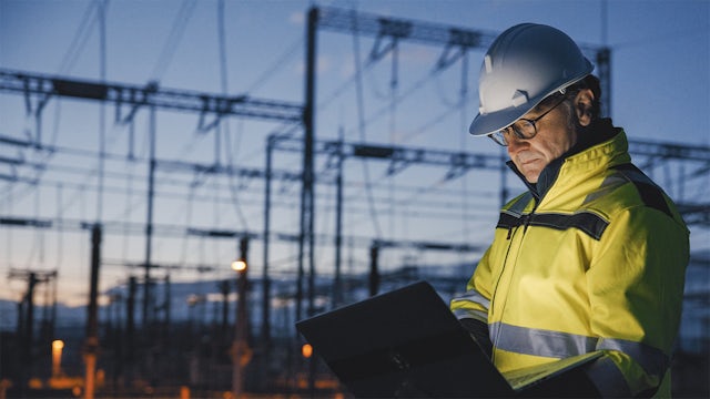 A worker in safety gear stands before a power plant, wearing a hard hat and a safety vest