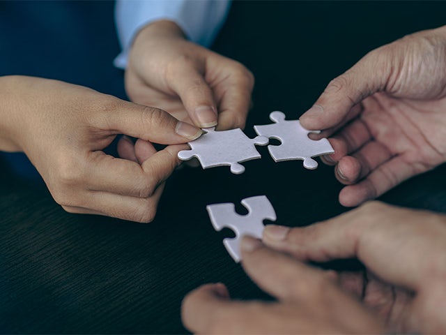 Three people's hands holding white puzzle pieces coming together