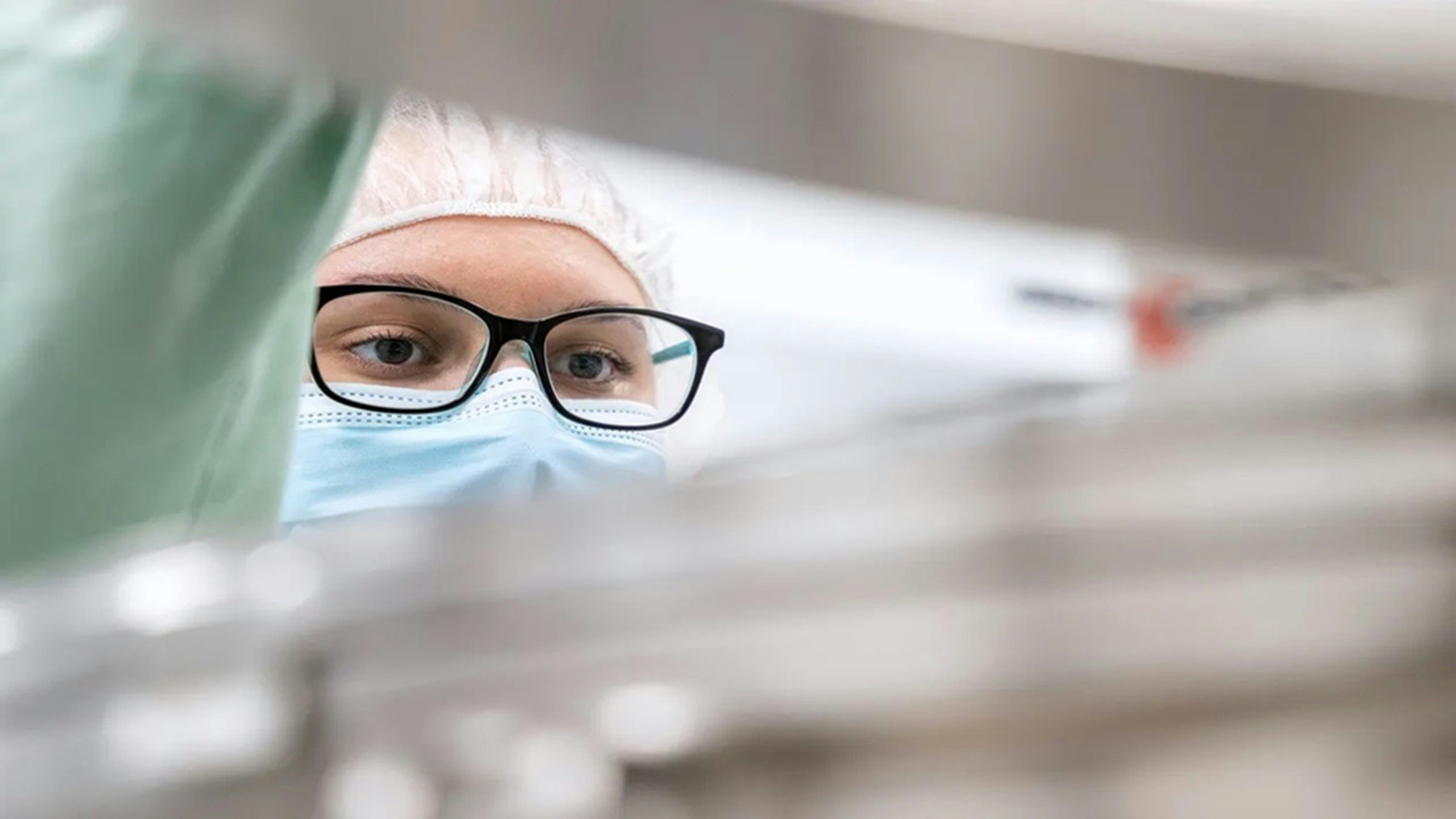 A person wearing a surgical mask, glasses and a hairnet looks intently upwards.