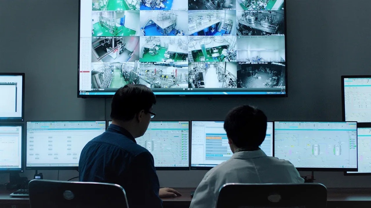 Two people monitoring a factory control room with multiple screens.