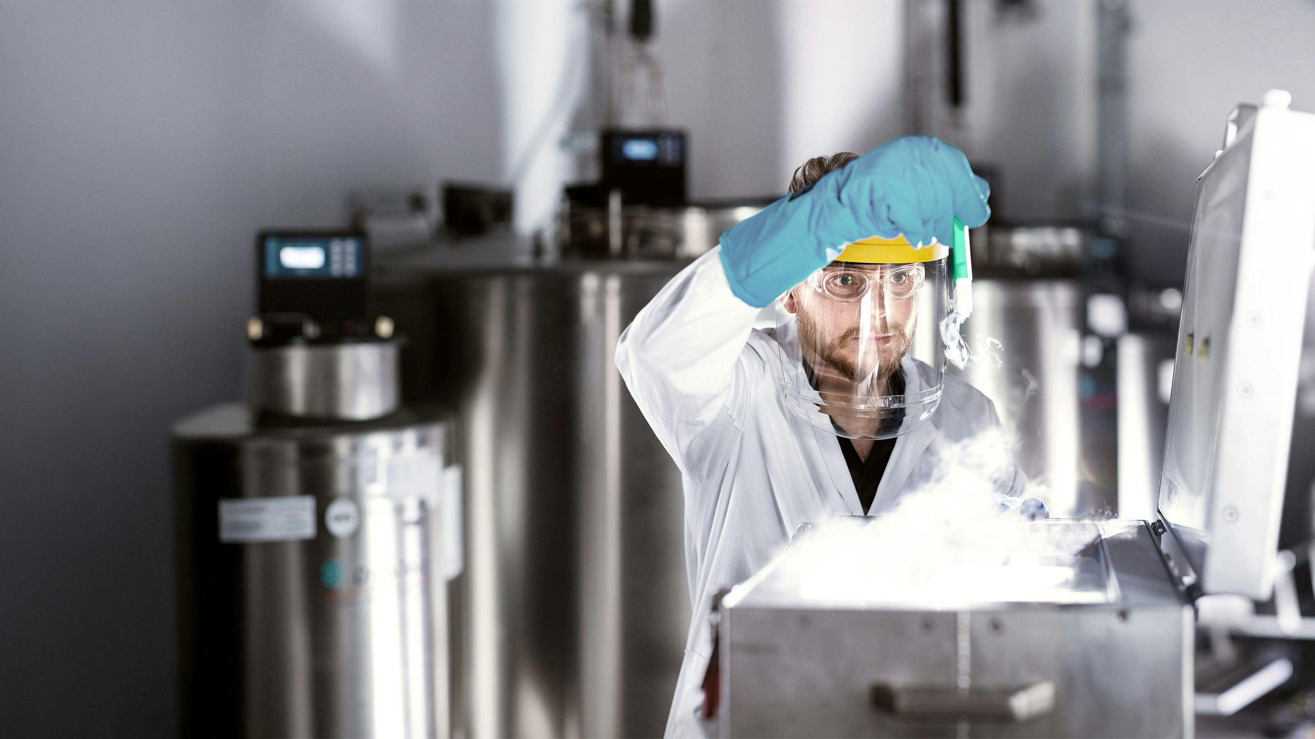 A bearded man wearing glasses and PPE, including blue gloves and a face mask, inspects an item in a pharmaceutical factory.