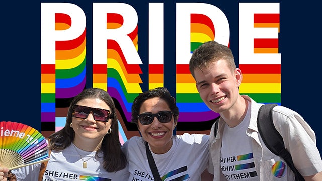 A group of people in a room, smiling and posing for a photo, with a rainbow flag in the background.