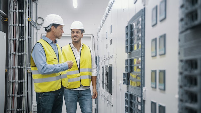 Two men in yellow vests and white hardhats converse in an industrial setting.