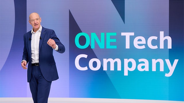 Man in a suit and tie speaking at a podium with a microphone and display screens in the background.