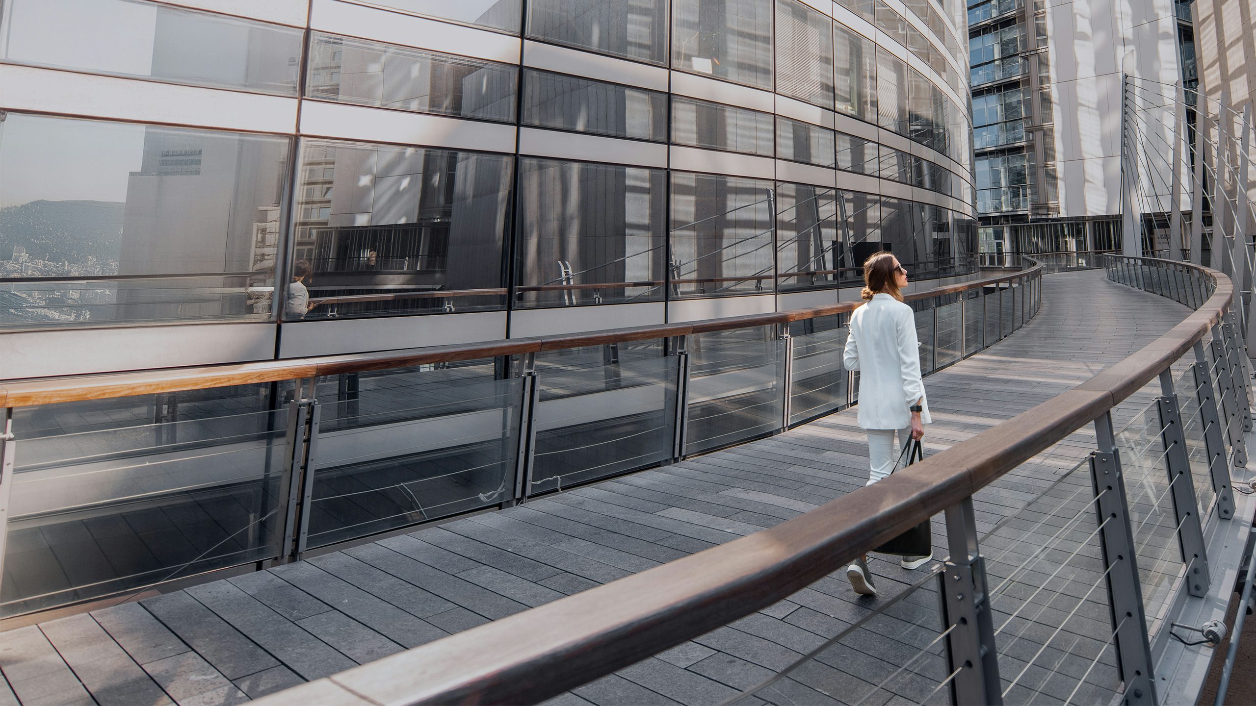 A woman in a white suit walks along a modern, curved outdoor walkway beside a glass-paneled building.