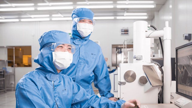 Two workers in blue cleanroom suits look at a computer monitor in a semiconductor factory.