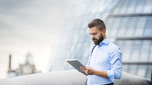 A man looking at information on a tablet device while standing on a walkway, with a building in the background.