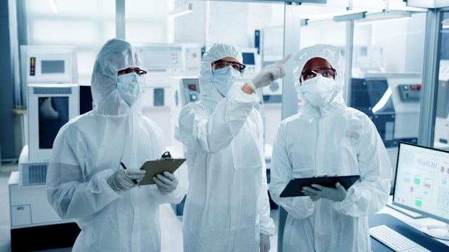 Three workers in white cleanroom suits and clipboards converse in a semiconductor factory.