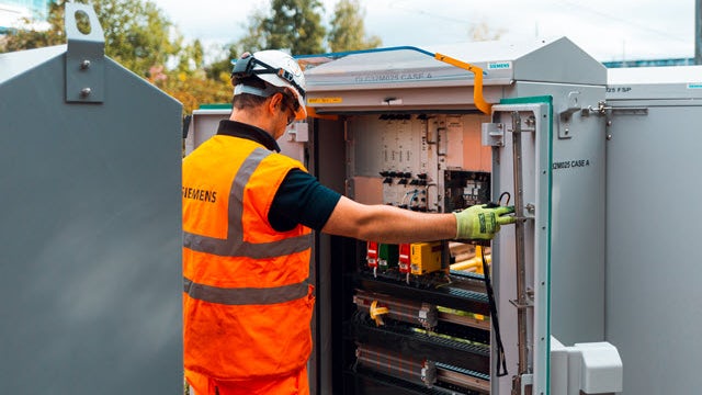 A person wearing a headset sits at a desk with multiple computer monitors displaying technical diagrams and data.