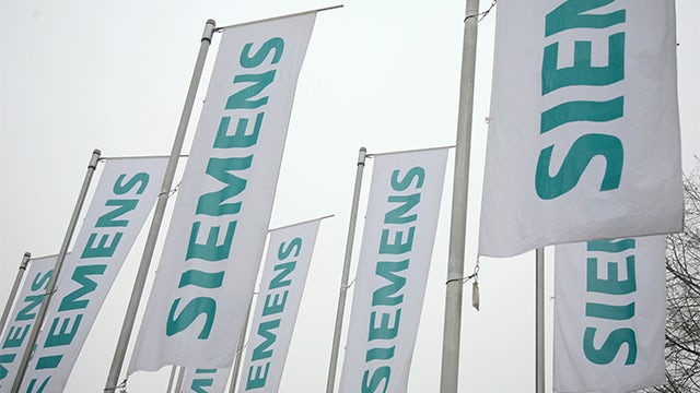 Several white flags with the Siemens logo in teal letters wave on tall flagpoles against a gray sky.