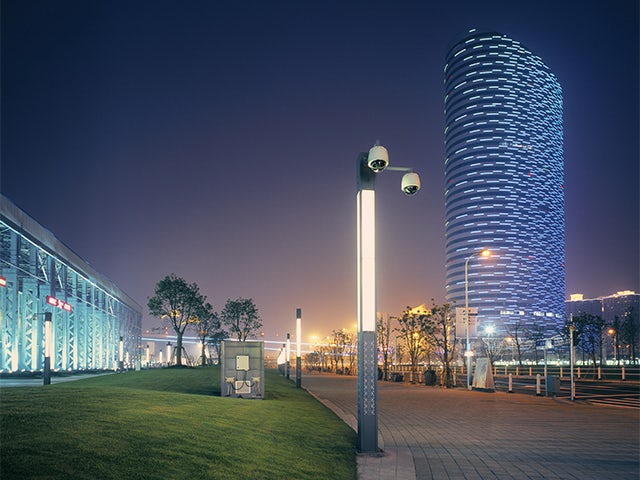 Modern city night scene with a tall, blue-lit skyscraper, a street light with security cameras, and a paved walkway.
