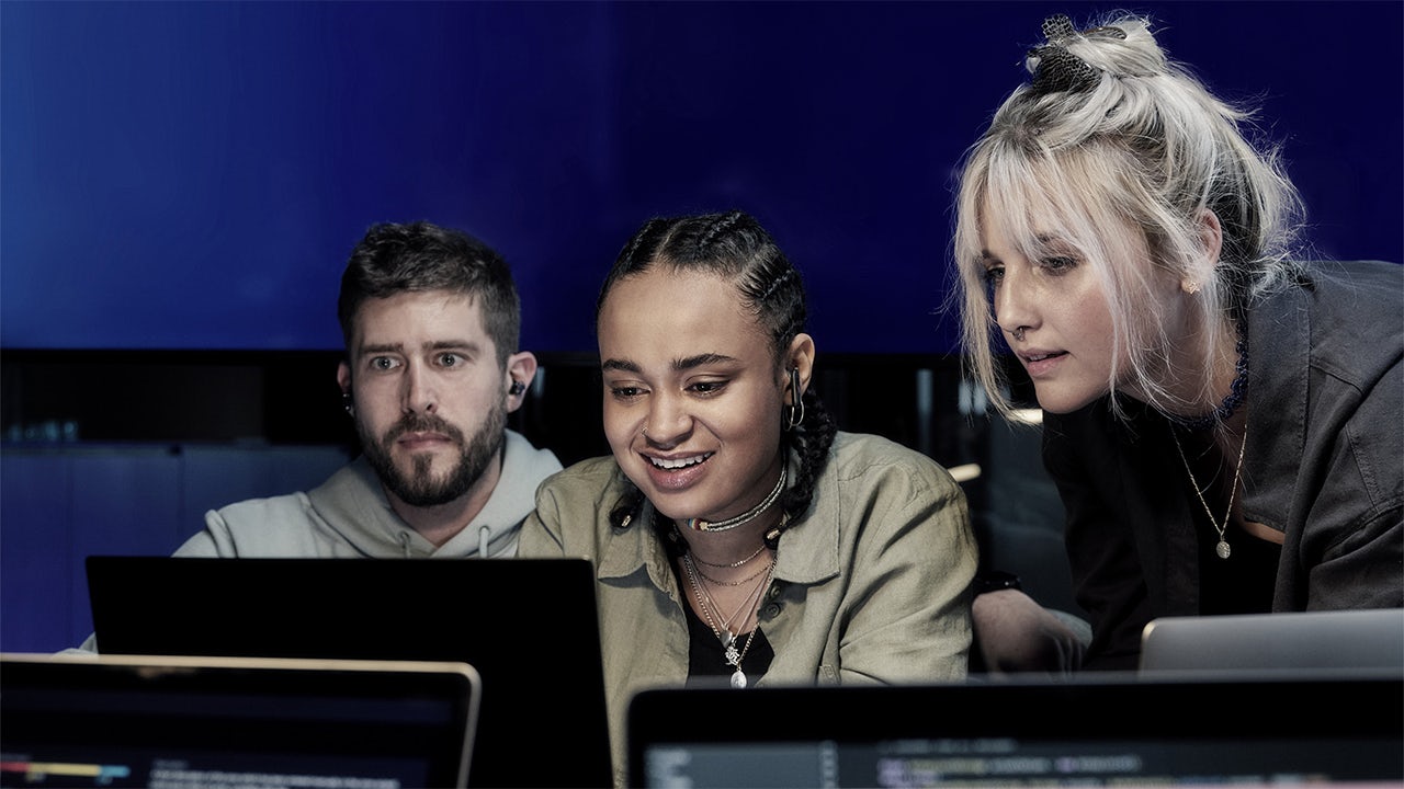 Three digital experts wearing headsets collaborate around a cluster of computers.