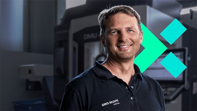 The Chief Product Officer of DMG MORI smiles in front of large manufacturing equipment alongside the Xcelerator logo.