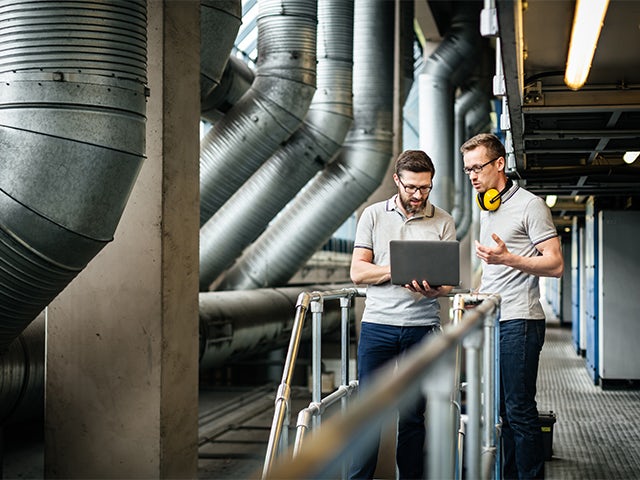 Two engineers with laptop standing in a large printery and discussing latest production data.