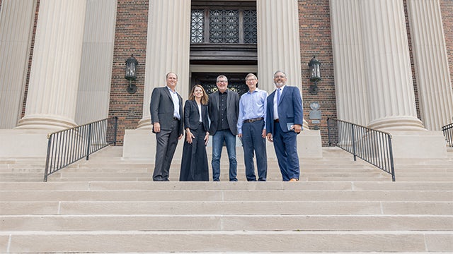 A group of five professionals, four men and one woman, in business attire standing on the steps of a university building.