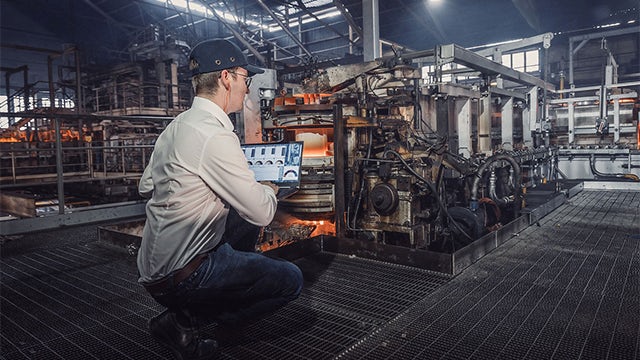 Man with laptop inspecting furnaces in a glass factory.