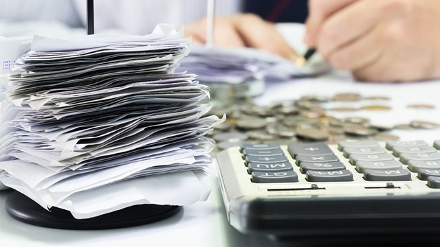 Closeup of calculator and pile of receipts with change and hands writing a check in the background.