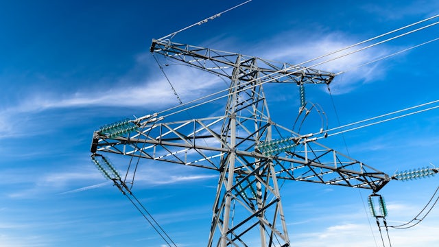 High-voltage electrical transmission tower with power lines and glass insulators against bright blue sky with clouds.
