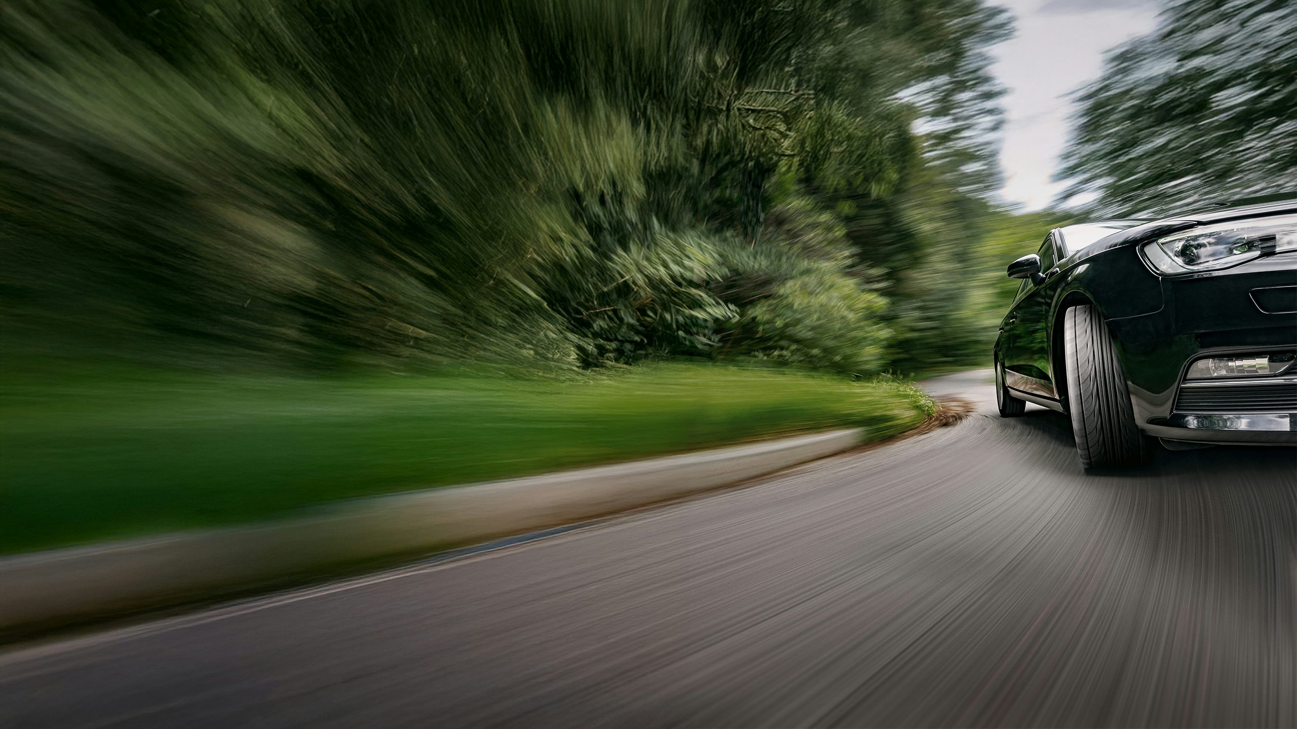 Prominently angled front tire of a black car rounding the right-hand bend on a blurred, tree-lined road, conveying speed.