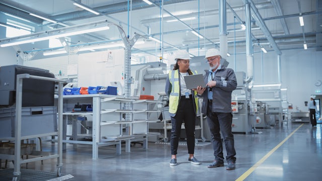 Two engineers in hard hats standing in factory looking at a laptop.