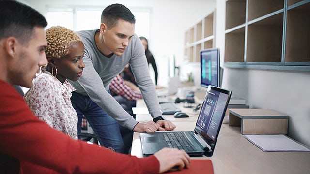 A team of professionals collaborates around a laptop, analyzing industrial automation data, illustrating IT/OT integration.