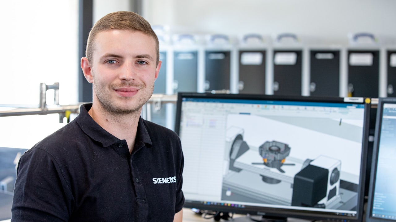 A Siemens employee stands in front of two computer monitors. 