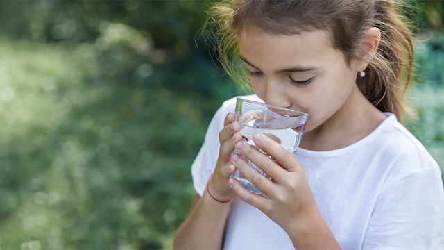 A little girl with a ponytail and a white T-shirt drinks water out of a clear glass.