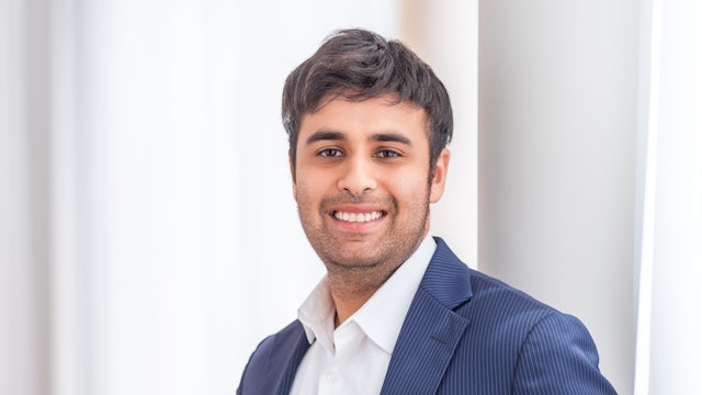 A person in a suit and tie, standing in front of a white wall with a black border, and appears to be posing for a photograph.