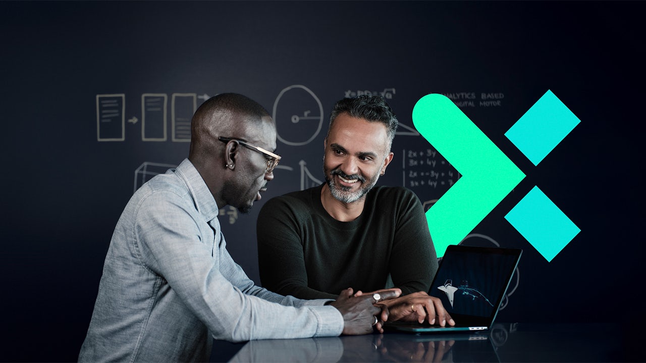 A person in a blue shirt smiles while sitting at a desk with a laptop and notebook in an office setting.