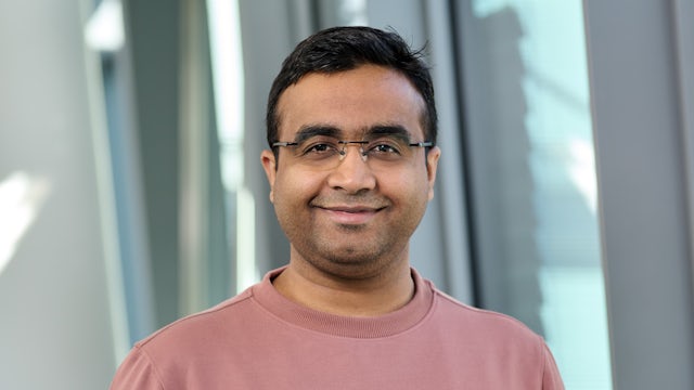 A person in a black shirt standing against a white wall, holding a dark object with a blurred background.