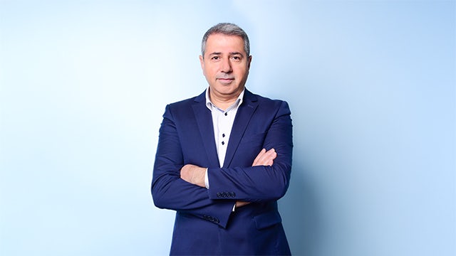 A man with a serious expression, wearing a suit and tie, is standing in front of a blurred background.