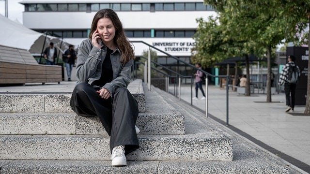Female student talks on phone while sitting on granite steps on the University of East London campus.
